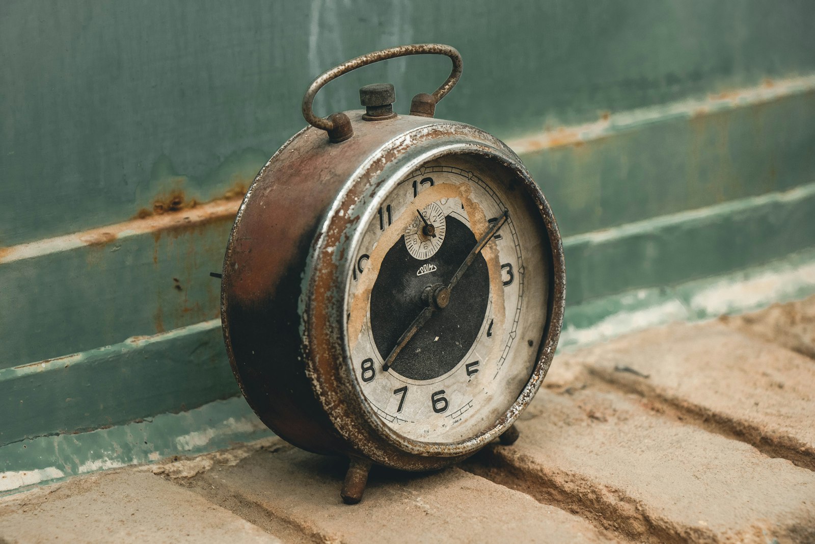 A rusted alarm clock propped against a peeling wall, hands still on the dial, long since stopped