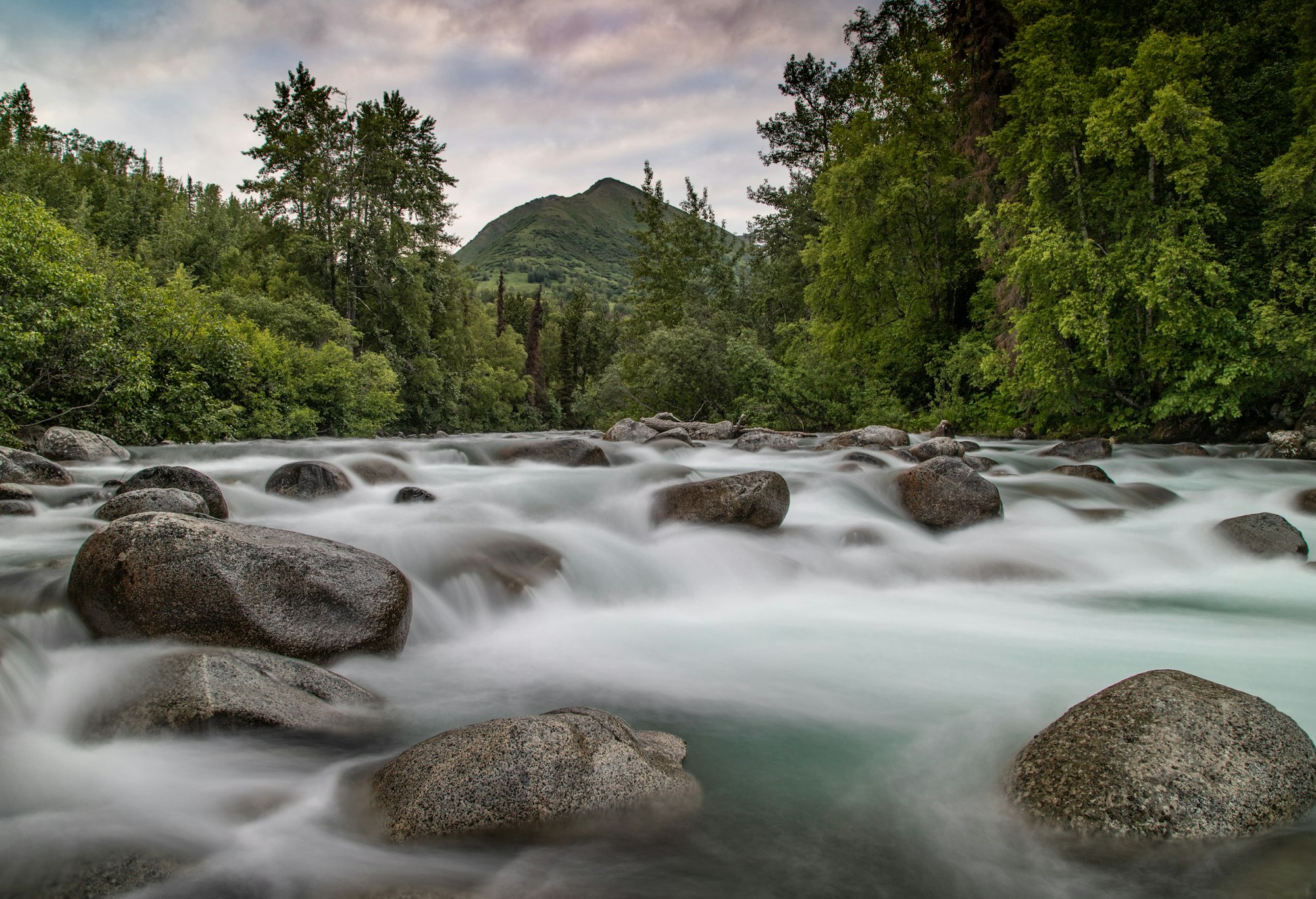 A river rushing over boulders between green wooded hills, the water blurred to a white haze by a long exposure